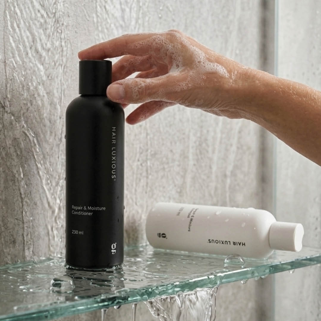 Hand reaching for the Hair Luxious Repair & Moisture Conditioner on a glass shower shelf