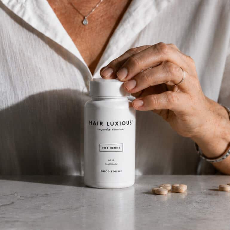 Hands opening a white Hair Luxious supplement jar on a marble counter, with tablets beside it